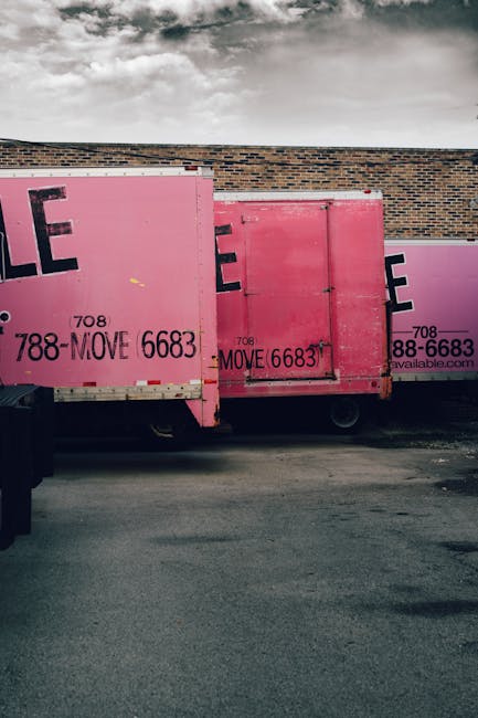 Two large pink moving containers with black text displaying contact information are positioned adjacent to a brick wall outdoors. The containers are mounted on a vehicle, which appears to be a truck, and are used for storing and transporting household furniture and packing materials during a home relocation. One container has a side door, which is closed, while the other is open, revealing cardboard boxes, plastic wrap, and protective blankets inside. The containers are situated on a paved area, likely a driveway or loading zone, with the sky overhead showing cloudy weather. Removal Companies Maida Vale may utilize such containers for efficient furniture transport and packing during house removals in Maida Vale. The environment is well-lit, with natural daylight illuminating the scene, emphasizing the practical process of loading packing supplies and furniture as part of the moving and unpacking logistics involved in a professional relocation service.