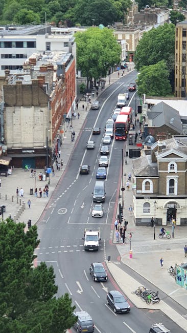 An aerial view capturing a busy street scene in Maida Vale with a line of vehicles, including cars and a double-decker bus, moving along the road. The street is lined with various buildings, including commercial and residential properties, with some featuring brick facades and others modern exteriors. Pedestrians are visible on the sidewalks, some waiting at a crosswalk and others walking past shops and houses. Green trees provide a canopy over parts of the street, adding a touch of greenery to the urban environment. In the foreground, bicycles are parked near a corner, while a person on a bicycle is crossing the road. The scene depicts typical city traffic and street activity, fitting within the context of home relocation and moving logistics services offered by Removal Companies Maida Vale, as they manage transport and moving processes within urban areas.