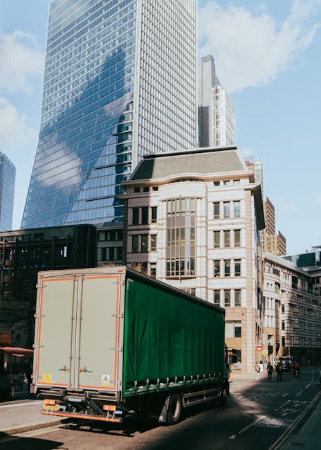 Two large pink moving containers with black text displaying contact information are positioned adjacent to a brick wall outdoors. The containers are mounted on a vehicle, which appears to be a truck, and are used for storing and transporting household furniture and packing materials during a home relocation. One container has a side door, which is closed, while the other is open, revealing cardboard boxes, plastic wrap, and protective blankets inside. The containers are situated on a paved area, likely a driveway or loading zone, with the sky overhead showing cloudy weather. Removal Companies Maida Vale may utilize such containers for efficient furniture transport and packing during house removals in Maida Vale. The environment is well-lit, with natural daylight illuminating the scene, emphasizing the practical process of loading packing supplies and furniture as part of the moving and unpacking logistics involved in a professional relocation service.