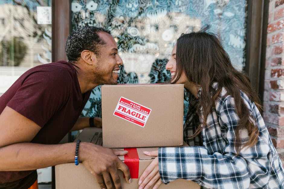 Two removal men are engaged in a furniture loading process at the rear of a white van on a paved street, with open rear doors revealing a large, green protective blanket covering a bulky item. One wor