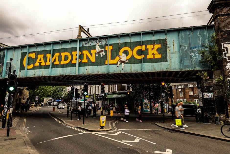 A large, weathered green metal railway bridge spanning a busy urban street in Maida Vale, London. The bridge displays a vivid yellow and red painted sign reading 'Camden Lock', with a small figure climbing on the letters. Beneath the bridge, pedestrians cross the street, some pushing strollers or carrying bags, while others wait at traffic lights. The street features multiple lanes, traffic signals, and vehicles including cars and a van. Various parcels, cardboard boxes, and wrapped furniture are visible on the pavement and near the curb, indicating ongoing home relocation and furniture transport activities. Removal Companies Maida Vale is involved in overseeing the loading process, with some items placed on trolleys or being carried towards a nearby van. The environment is urban, with brick buildings, street art, and signage nearby, illuminated by natural, slightly overcast daylight, supporting the context of packing and moving logistics in a busy city setting.