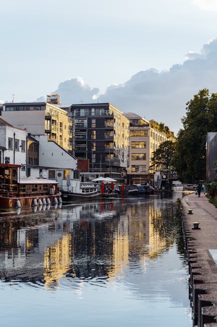 A view of a canal in Maida Vale, showing several residential modern apartment buildings with glass balconies and contemporary architectural features lining the water's edge. The canal is calm, reflecting the buildings and a partly cloudy sky overhead. On the right side of the image, a paved walkway runs alongside the canal, separated by a low stone or concrete edge, with some greenery and trees extending beyond the walkway. In the foreground, a small boat is docked close to the shore, and a few people can be seen walking along the pavement. The scene suggests a quiet, urban environment during daylight hours, with natural light illuminating the buildings and water surface, highlighting the proximity of residential properties to water, which is relevant to the relocation and home moving services offered by Removal Companies Maida Vale.