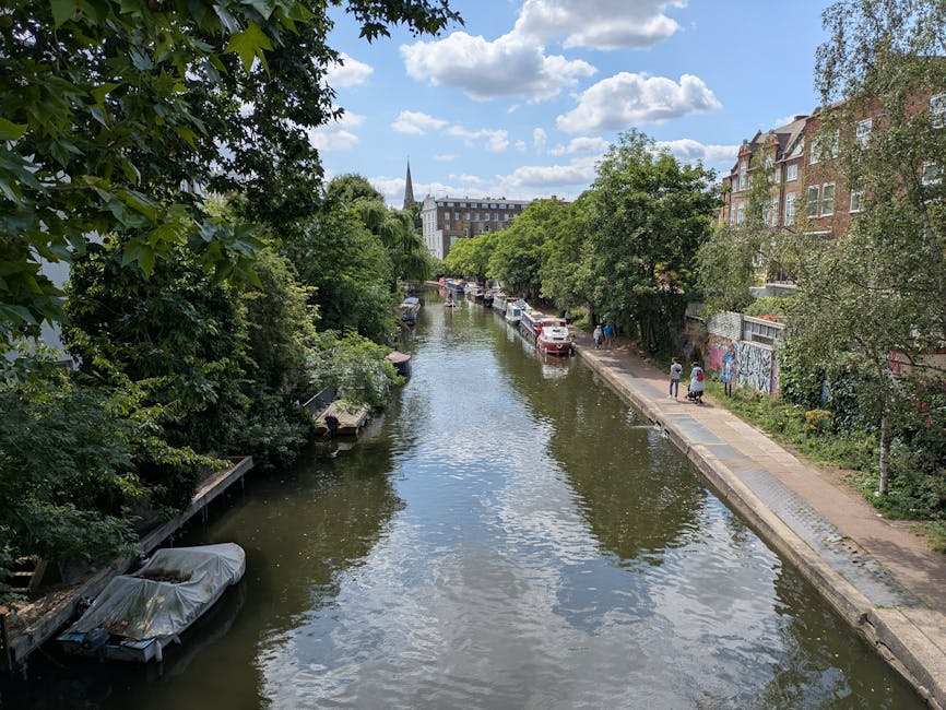 A canal scene in Maida Vale featuring calm water reflecting partly cloudy skies, with several houseboats and boats moored along the banks. The towpath on one side shows a few pedestrians walking, while on the opposite side, parked cars are visible next to a row of brick residential buildings with trees partially shading the area. The image captures a peaceful urban setting typical of the area, with greenery lining the canal and a church steeple visible in the distance. This scene relates to house removals or logistics services offered by Removal Companies Maida Vale, highlighting the environment where moving and furniture transport operations may take place within this picturesque waterway setting.