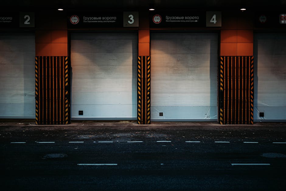 A row of three large, closed warehouse or loading bay doors with white panels, each framed by black and yellow striped safety barriers. Above the doors are numbered signs labeled '2,' '3,' and '4,' with additional signage in Russian or Cyrillic script. The scene is outdoors, in front of a paved street with white road markings, and the area appears dimly lit, suggesting early morning or evening. The setting indicates a facility where furniture transport or home relocation operations might occur, with the presence of loading bays designed for vehicle access, supporting the logistics involved in removals and packing processes, as managed by Removal Companies Maida Vale.