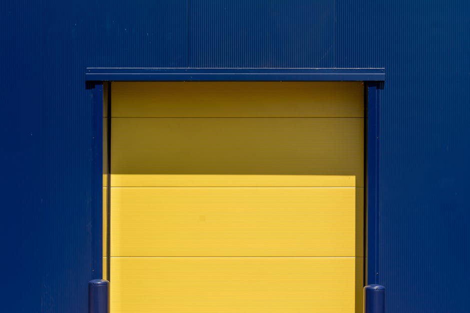 A large yellow industrial roll-up door set within a blue metal building exterior, with two blue bollards positioned on either side of the doorway. The door appears to be closed, and the surrounding environment suggests an outdoor loading bay area used for home relocation or furniture transport services. This scene is associated with the process of loading or unloading items during a house removal, with the blue metal panels providing a sturdy backdrop for moving activities. The lighting highlights the contrasting colours of yellow and blue, emphasizing the industrial setting where Removal Companies Maida Vale might conduct their moving operations in compliance with local council rules for loading bay permits, as indicated on the webpage about house removals in Maida Vale.