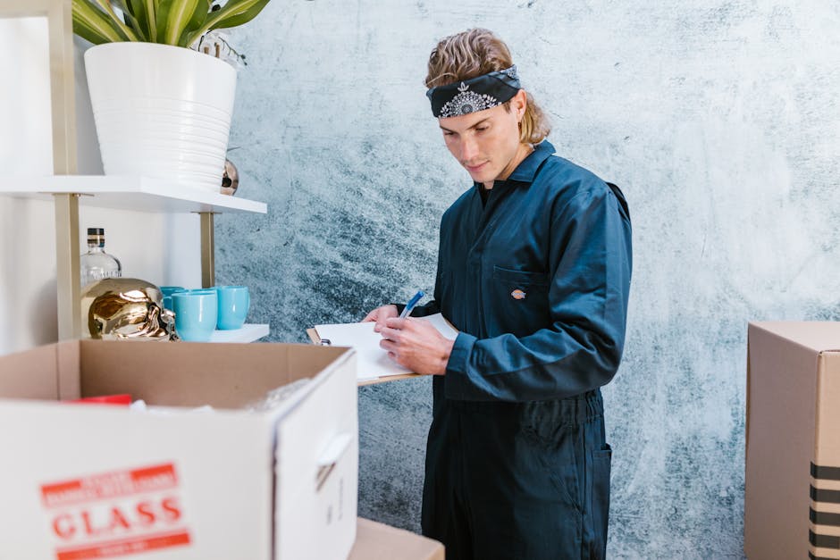 A young man wearing a black bandana and a dark blue work jumpsuit stands inside a room with a textured blue wall. He is holding a clipboard and pen, appearing to take notes during a house removal or packing process. In front of him, there are open cardboard boxes filled with items, including glassware and ceramic mugs. To his left, there are white shelving units with decorative objects, such as a gold-colored sculpture, bottles, and blue cups. The environment is well-lit, and the scene captures a moment of organisation during furniture transport or home relocation, with the professional from Removal Companies Maida Vale preparing logistics for a moving services task.