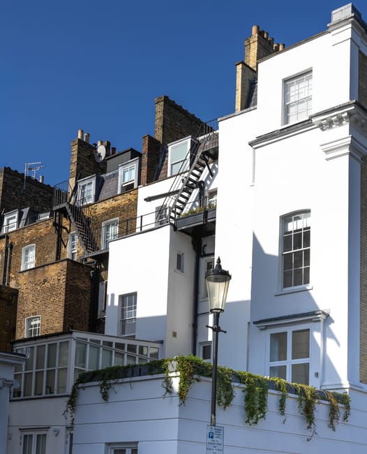 A residential street scene showing a multi-storey UK house with a white facade and large windows, adjacent to older brick-built buildings with chimneys and external fire escape stairs. The house features a small balcony with a glass-enclosed conservatory on the lower level, and a black wrought iron street lamp stands near the front. The sky is clear and blue. This image is relevant to house removals and home relocation services offered by Removal Companies Maida Vale, illustrating typical urban architecture in Maida Vale, where furniture transport and packing may be necessary during moves.