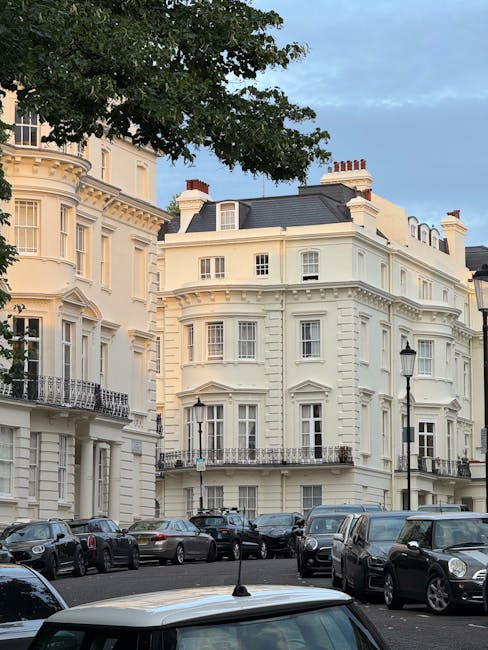 A street view outside a large, cream-colored Victorian-style building in Maida Vale, NW6, with multiple floors, bay windows, and decorative architectural details. Several parked cars line the street, including sedans and SUVs, in front of the property. Tall black lampposts are positioned along the pavement, and a large leafy tree partially shades the scene. Inside the property, visible through open doors, are cardboard boxes, furniture wrapped in protective fabric and plastic, and a hand truck, indicating an ongoing home relocation or furniture transport process. The environment suggests day-time, with natural light illuminating the building and street, and the scene is associated with professional removals services provided by Removal Companies Maida Vale during packing and moving activities.