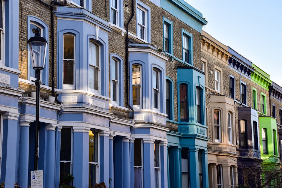 A row of multi-storey Victorian terraced houses in Maida Vale with colorful bay windows and decorative architectural details, situated along a pavement with a classic black street lamp in the foreground. The buildings feature a variety of pastel shades, including light blue, teal, beige, and lime green, with some windows reflecting the sky and surroundings. The scene is captured in daylight with clear weather, highlighting the urban residential environment. This image relates to house removals and moving services provided by Removal Companies Maida Vale, illustrating the type of property involved in home relocation and furniture transport processes.