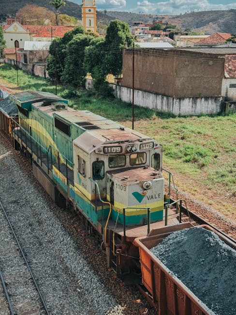 A freight train with a weathered cream, green, and blue locomotive numbered 1109 is situated on railway tracks in an urban environment. The locomotive features a rounded front with multiple headlights and a small windshield, and is connected to a series of freight cars, including a wagon loaded with dark gravel or crushed stone. The train runs through a semi-rural area with green grass, trees, and a few residential or industrial buildings in the background, including a yellow clock tower nearby. The scene is captured in daylight with clear visibility, and the train appears to be in motion or about to begin a loading process related to logistics and transportation. The image is relevant for illustrating aspects of home relocation logistics involving furniture transport by train, or the movement of goods during a house removal, consistent with services offered by Removal Companies Maida Vale.