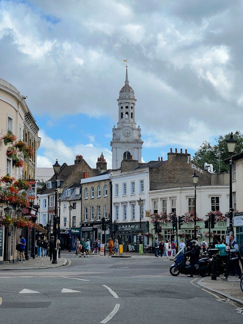 A busy street scene in Maida Vale with a historic white clock tower and spire dominating the skyline under a partly cloudy sky. In the foreground, a row of buildings featuring various shops and cafes, some with hanging flower baskets, line the street. Pedestrians are seen walking along the pavement, some carrying shopping bags, while others stand near the shops. To the right, a black scooter is parked on the pavement, and a small van is partially visible, possibly involved in a home relocation or moving process. The road shows marked lanes, and a few vehicles are passing by or parked nearby. The scene captures a lively environment typical of a central urban area suitable for house removals and furniture transport services provided by Removal Companies Maida Vale, with the environment reflecting a mix of commercial activity and daily life in the neighbourhood.
