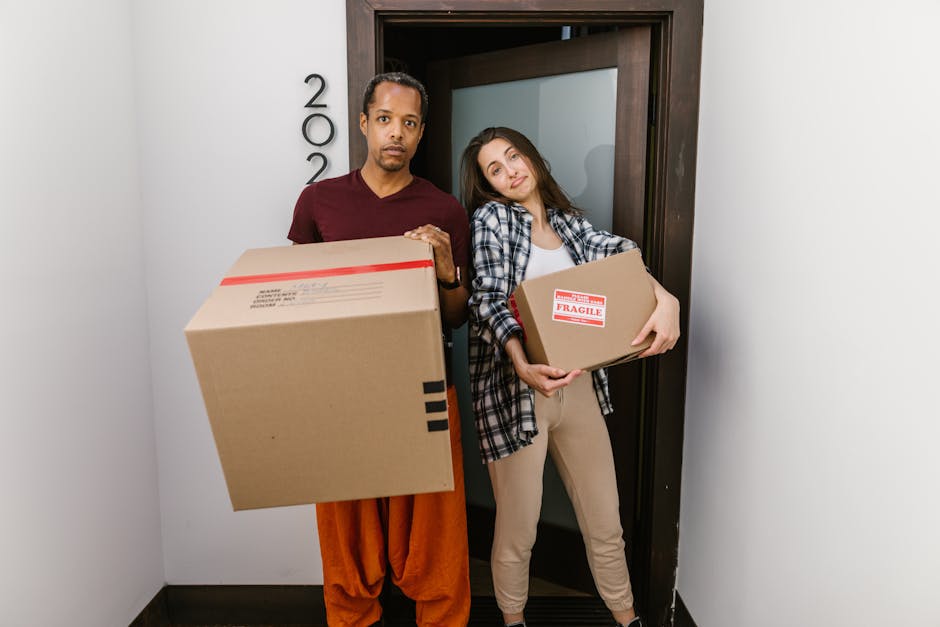 A man and woman stand inside a residential building near a doorway, each holding cardboard boxes for home relocation. The man, with short dark hair and wearing a maroon t-shirt and orange pants, holds a large box with black markings, while the woman, with long brown hair and dressed in a plaid shirt and beige trousers, holds a smaller box labeled 'Fragile' with red tape. The scene shows an indoor setting with plain white walls, a dark door frame, and a glimpse of the hallway behind them. The individuals appear to be preparing for a furniture transport or packing and moving process, guided by the services of Removal Companies Maida Vale. The lighting is even and natural, emphasizing the preparation involved in house removals, packing fragile items, and loading boxes for transport.