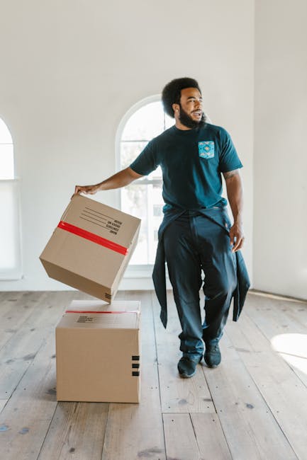 A man with a dark afro hairstyle and a beard is inside a bright, minimally furnished room with large arched windows allowing natural light to fill the space. He is wearing a navy blue T-shirt and dark trousers, holding a cardboard box in his right hand, which he is in the process of placing onto a second box on the light wooden floor. The boxes are sealed with packing tape, indicating they are packed for a home relocation. The room’s neutral walls and the clean, open environment suggest a move-in or move-out scenario managed by a professional removal service. There are no visible furniture or appliances in the image, emphasizing the packing and moving process, with the man appearing to coordinate or organize the loading of boxes. This scene reflects components of furniture transport and packing for house removals, relevant to services such as those offered by Removal Companies Maida Vale.