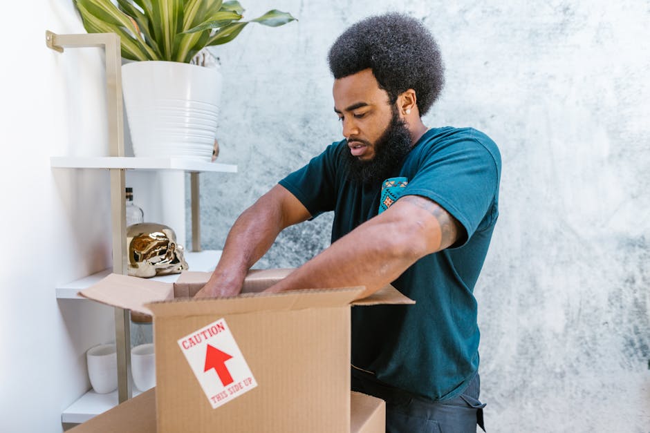 A man and woman stand inside a residential building near a doorway, each holding cardboard boxes for home relocation. The man, with short dark hair and wearing a maroon t-shirt and orange pants, holds a large box with black markings, while the woman, with long brown hair and dressed in a plaid shirt and beige trousers, holds a smaller box labeled 'Fragile' with red tape. The scene shows an indoor setting with plain white walls, a dark door frame, and a glimpse of the hallway behind them. The individuals appear to be preparing for a furniture transport or packing and moving process, guided by the services of Removal Companies Maida Vale. The lighting is even and natural, emphasizing the preparation involved in house removals, packing fragile items, and loading boxes for transport.