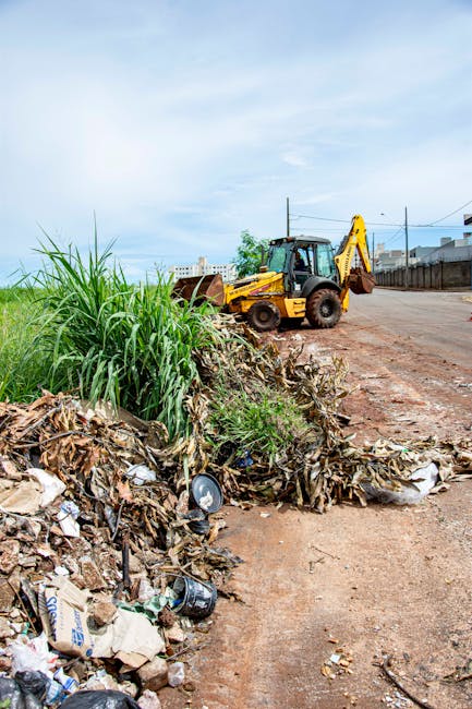 A yellow backhoe loader with a black hydraulic arm is positioned on a dirt roadside in an urban area during daytime, near a pile of mixed waste including cardboard, plastic, and organic debris alongside some overgrown grass. The loader appears to be in the process of clearing or transporting waste from the site, with the large bucket resting on the ground. In the background, there is a metal fence, utility poles, and a row of buildings under a partly cloudy sky. The scene suggests activity related to waste disposal or site clearing, which may be part of a property cleanup prior to or following a house removal or renovation project. The environment appears somewhat dusty, with the vehicle and waste pile being the main focal points, and the overall scene situated within an area where removal services, such as those offered by Removal Companies Maida Vale, may assist with logistics and site clearance tasks during home relocations or property preparations.