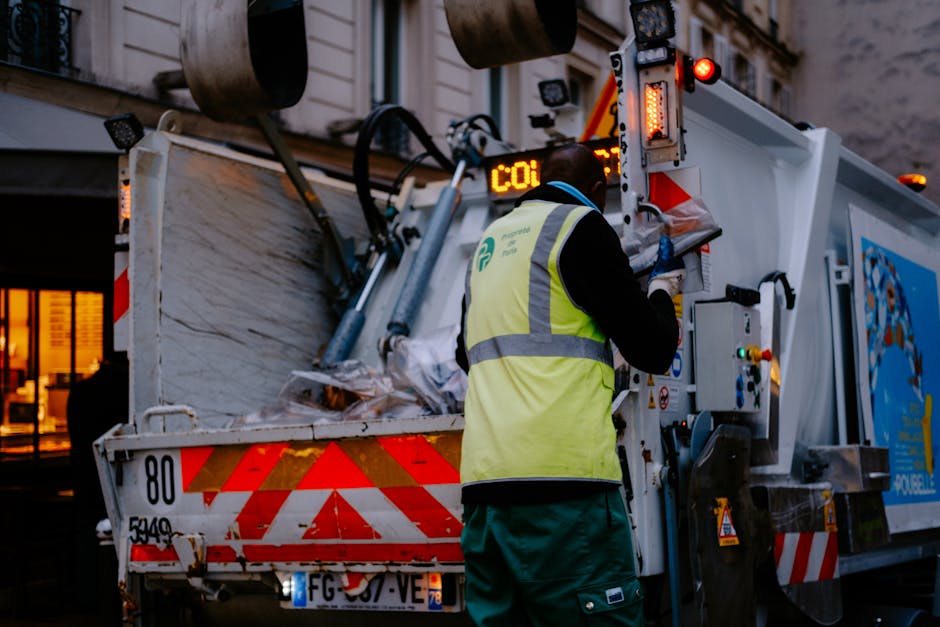 A sanitation worker wearing a high-visibility yellow vest and dark clothing operates a waste collection truck on a street during dusk or early evening. The worker is standing beside the vehicle, which is equipped with various mechanisms and warning lights, and appears to be managing the loading process of waste materials, possibly including cardboard boxes, plastic bags, and other packaging, from the ground into the truck's rear compartment. The truck is marked with red and white reflective safety stripes and has an open rear hatch filled with collected refuse. In the background, there is a building with brightly lit windows, indicating an urban environment. The image captures the process of waste disposal, an essential part of home relocation and moving logistics, supported by professional removal services such as those offered by Removal Companies Maida Vale.