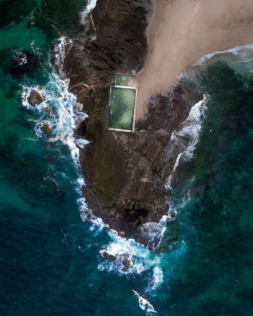 Aerial view of a coastal scene showing a small sandy beach and rugged rocky cliffs. There is a rectangular swimming pool filled with greenish water, situated on a flat, concrete platform near the shoreline. The seabed has varying shades of blue and turquoise, with waves crashing against the rocks and shoreline. The image captures the contrast between the natural landscape and the man-made pool, with some vegetation visible on the cliffs. The scene appears calm and isolated, with no people or boats in sight, illustrating a remote seaside location potentially relevant for house removals or relocation logistics near a coastal area.