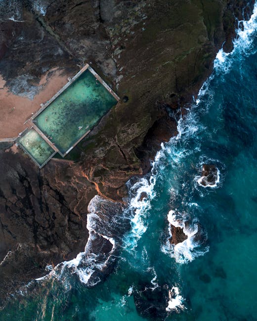 Aerial view of a coastal scene showing a small sandy beach and rugged rocky cliffs. There is a rectangular swimming pool filled with greenish water, situated on a flat, concrete platform near the shoreline. The seabed has varying shades of blue and turquoise, with waves crashing against the rocks and shoreline. The image captures the contrast between the natural landscape and the man-made pool, with some vegetation visible on the cliffs. The scene appears calm and isolated, with no people or boats in sight, illustrating a remote seaside location potentially relevant for house removals or relocation logistics near a coastal area.