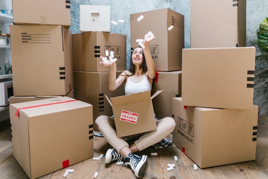 A man and a woman standing inside a residential building near the doorway, holding cardboard boxes used for packing and moving. The man wears a maroon T-shirt and orange trousers, carrying a large box with shipping labels, while the woman, dressed in a checkered shirt and beige pants, holds a smaller box marked 'Fragile.' Behind them, the door frame is visible, leading into an interior space, with a neutral wall on one side displaying room numbers. The scene captures the loading process during a home relocation, with both individuals preparing to move furniture, boxes, and household items, possibly as part of a professional removals service by Removal Companies Maida Vale. The environment appears well-lit, emphasizing the packing materials, the positioning of the boxes, and the preparation for transport outside or inside the property.